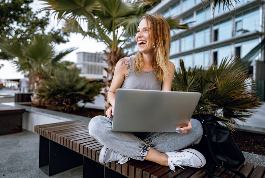A happy woman with strawberry blonde hair is looking off camera and laughing. She is sitting cross-legged on a bench outside and has a laptop on her lap. There is a cool tattoo of a flower on her right shoulder.