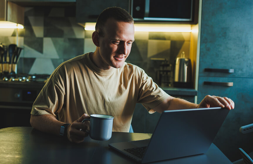A man with a crew cut is sitting at a kitchen table. He is opening a laptop to browse Usenet with his Newshosting account.