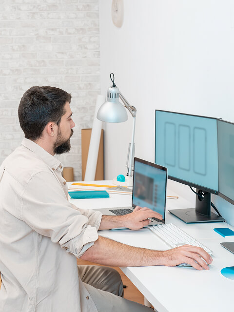 A man in a well-lit room is working on a desktop and a laptop at a white desk.