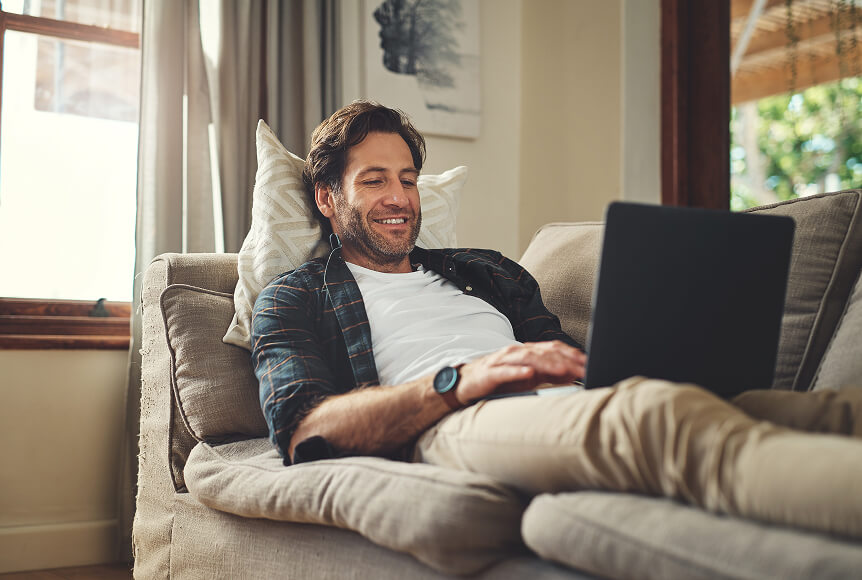 A man with a stubbly beard is laying back on a couch and smiling as he browses Usenet on his laptop.