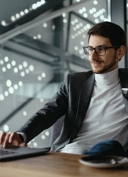 A man wearing a white turtleneck and sport coat is sitting at a conference table holding a black tea cup in his right hand while typing with his left hand on a laptop.
