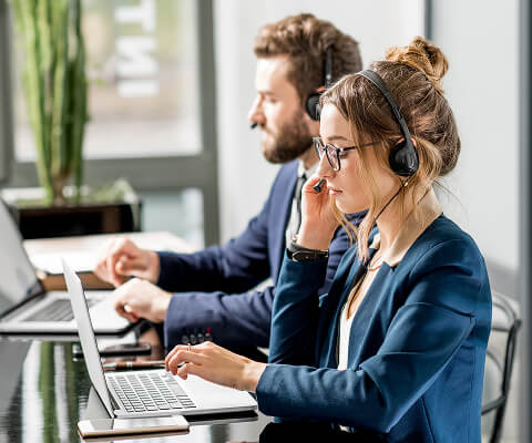 Two support team members are sitting at a glass-topped desk with laptops. There is a man with a beard and a blue jacket in the background and a woman with her hair in a bun and a blue blazer in the foreground. Both are wearing headsets, presumably to help Newshosting customers.