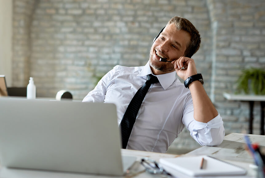 A man with short hair and wearing a shirt and tie is leaning back, smiling and talking on a headset.