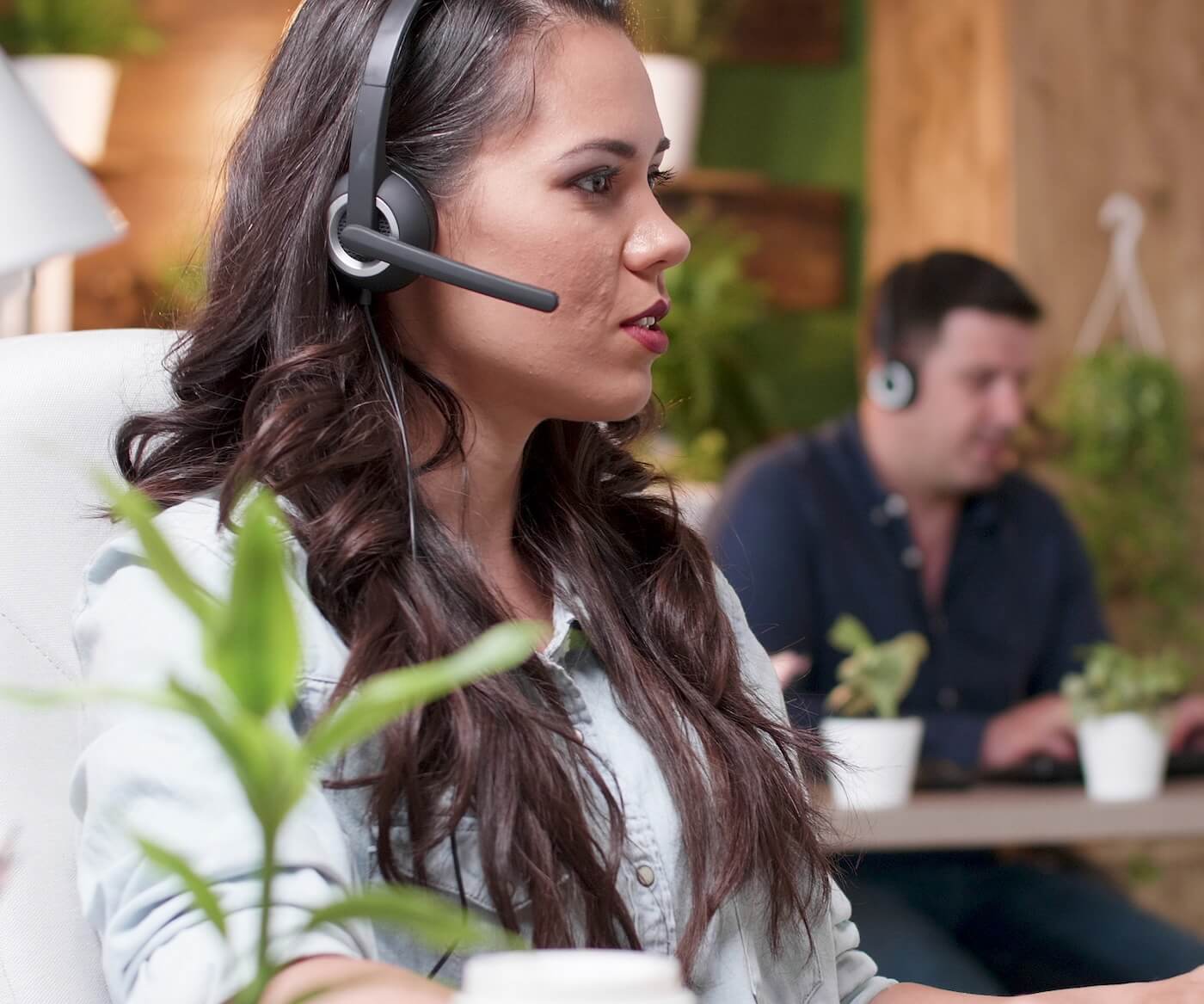 A woman is sitting at a desk with plants on it and helping Newshosting customers over a headset. There is a man at a similar desk doing the same in the background.