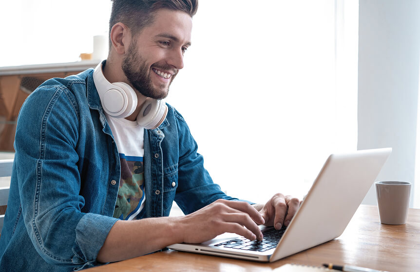 A man wearing a jean jacket, white headphones around his neck, and a graphic t-shirt is typing on a laptop and smiling because he is browsing Usenet with Newshosting, the best, fastest, most complete Usenet provider.