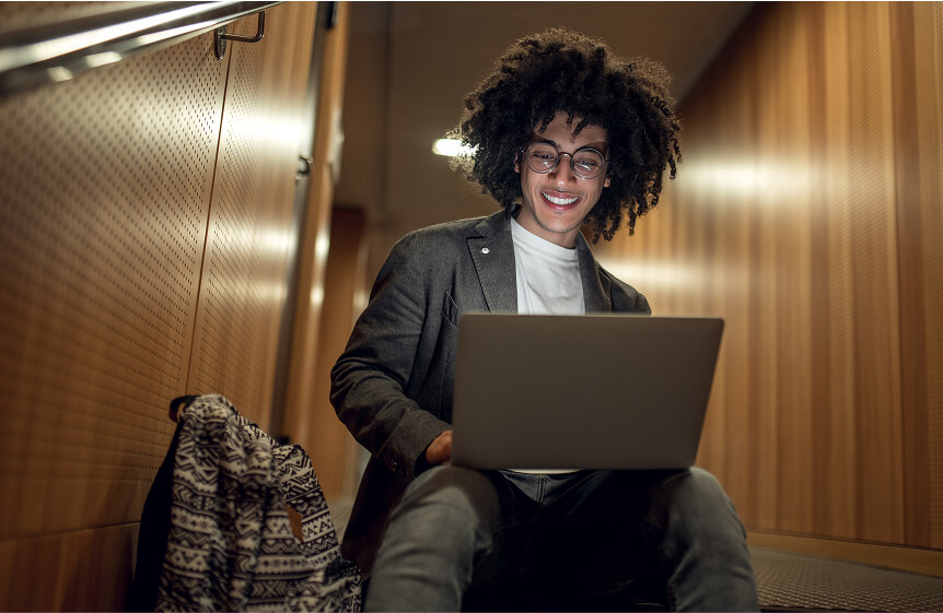 A person with amazing hair, just a huge, voluminous head of brown finger coils, great round glasses, and a fabulous canvas coat is sitting on a flight of stairs on their laptop. They are browsing Usenet on a laptop and smiling wide.