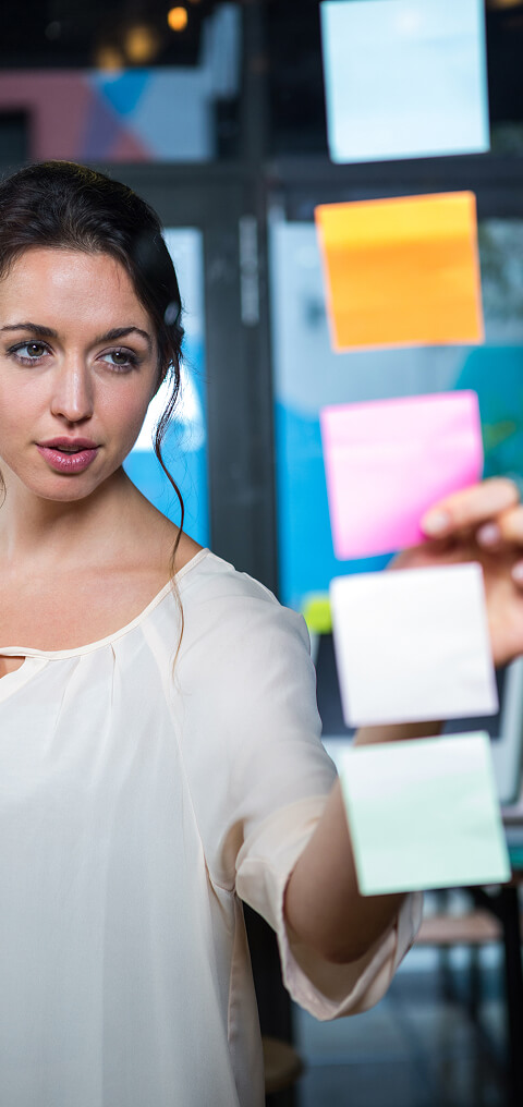 A woman in a white shirt is organizing her thoughts with sticky notes on a glass wall between the camera and her.