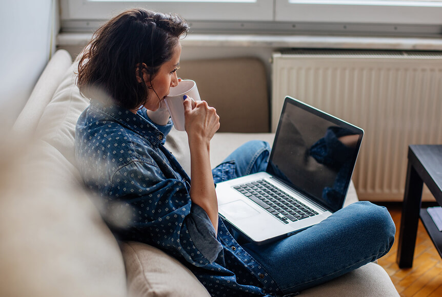 A woman with shoulder length hair wearing a blue polka-dotted shirt and jeans is waiting for her laptop to boot up while she drinks a cup of coffee.