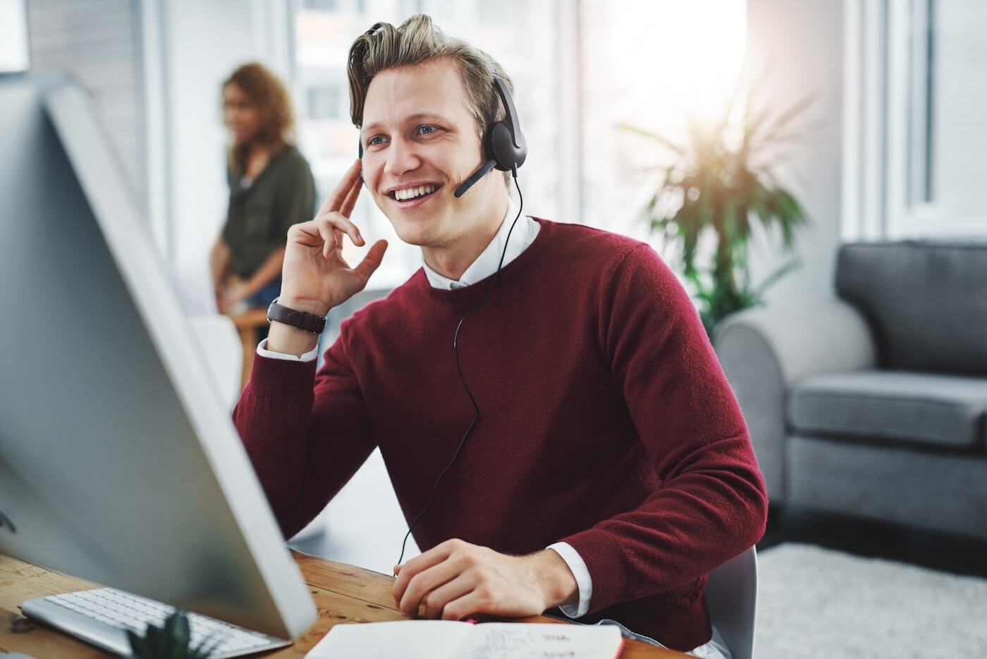 A man in a red sweater over a white buttondown shirt. He is smiling and talking to somebody on a headset while sitting in front of a desktop.