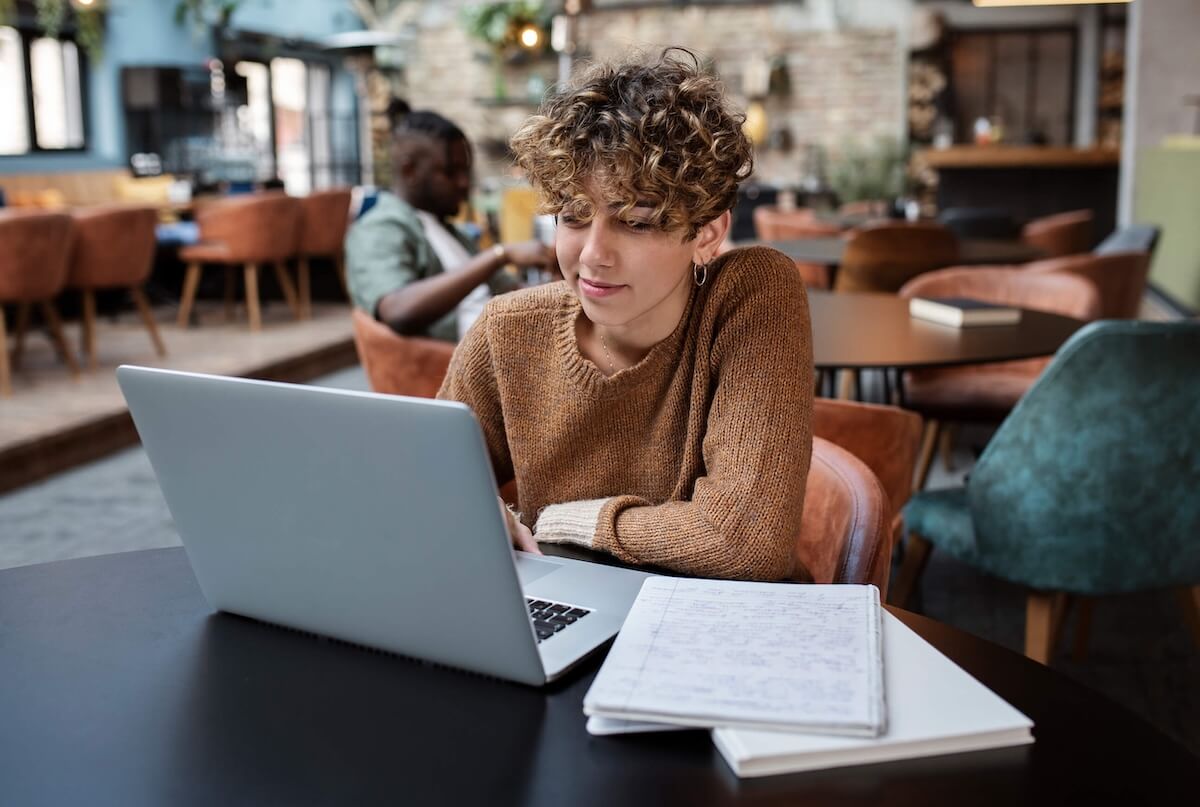A woman with short, curly hair wearing a brown sweater is browsing newsgroups on a laptop in a café.