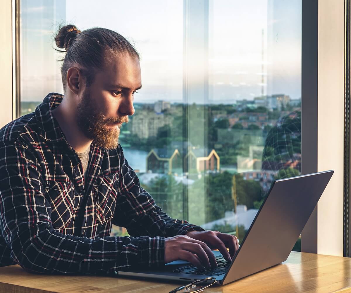 A man with his hair in a bun is sitting at a desk, typing on a laptop. There is a pastoral, small town in the window behind him.