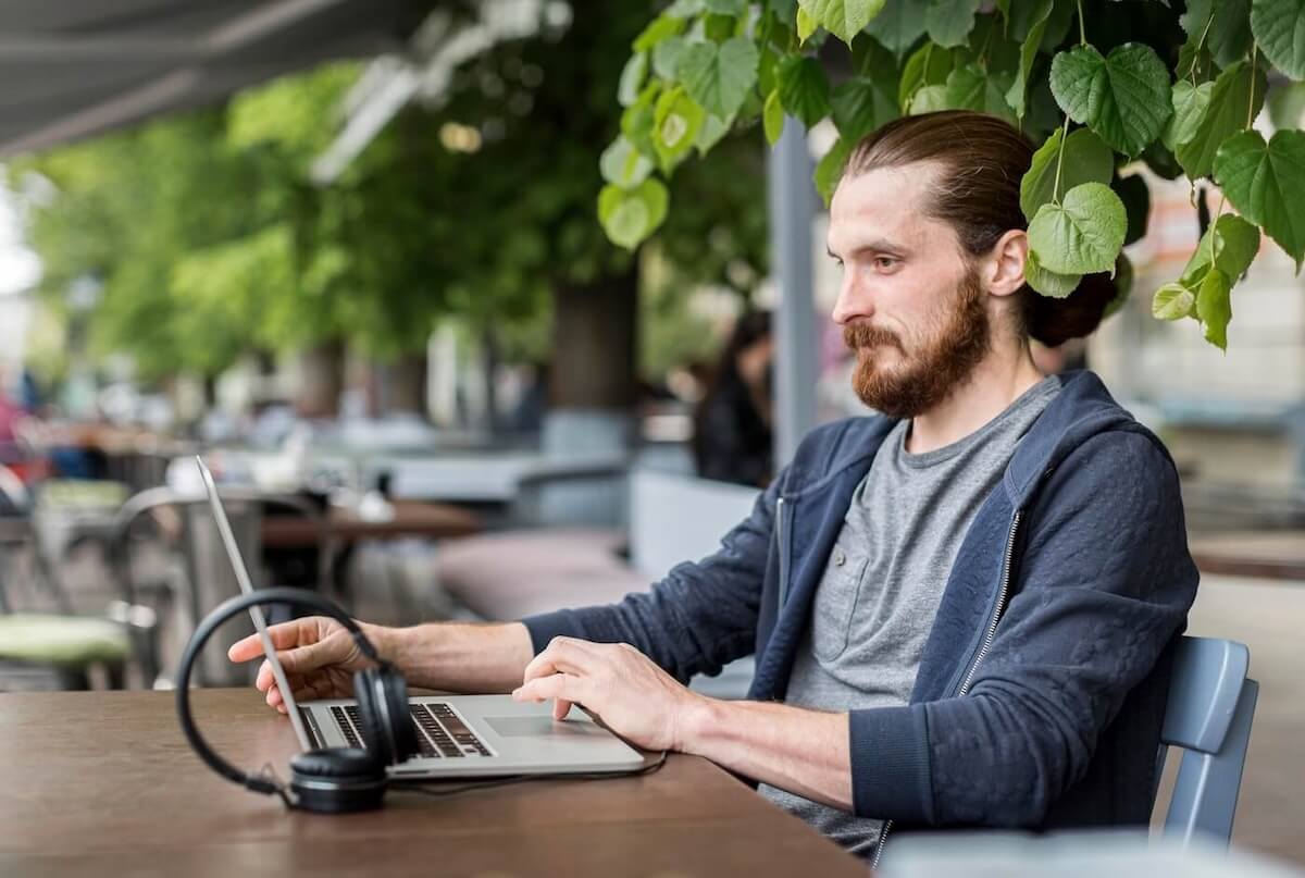 A man is sitting at a table outside. There is a tree growing behind his head and he is securely browsing on his laptop using a VPN.